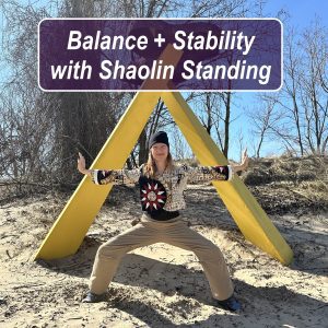 Melissa doing Shaolin standing pose outdoors beneath a yellow triangle structure, demonstrating balance and stability with grounded stance and outstretched arms in a natural sandy landscape.